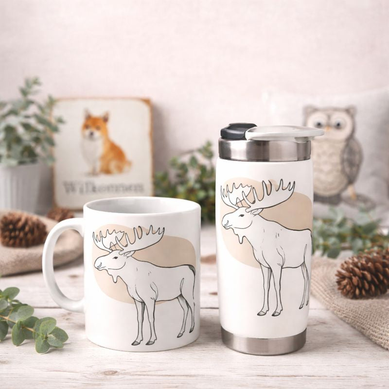 Keramikbecher und Thermobecher mit Elchmotiv auf heller Holzplatte vor sandfarbener Wand, mit Eukalyptus und Deko im Hintergrund.