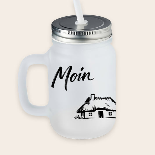 Satiniertes, matt-weißes Trinkglas mit Henkel, Metalldeckel und weißem Strohhalm, bedruckt mit einem schwarzen Reetdachhaus-Motiv und dem Schriftzug „Moin“.